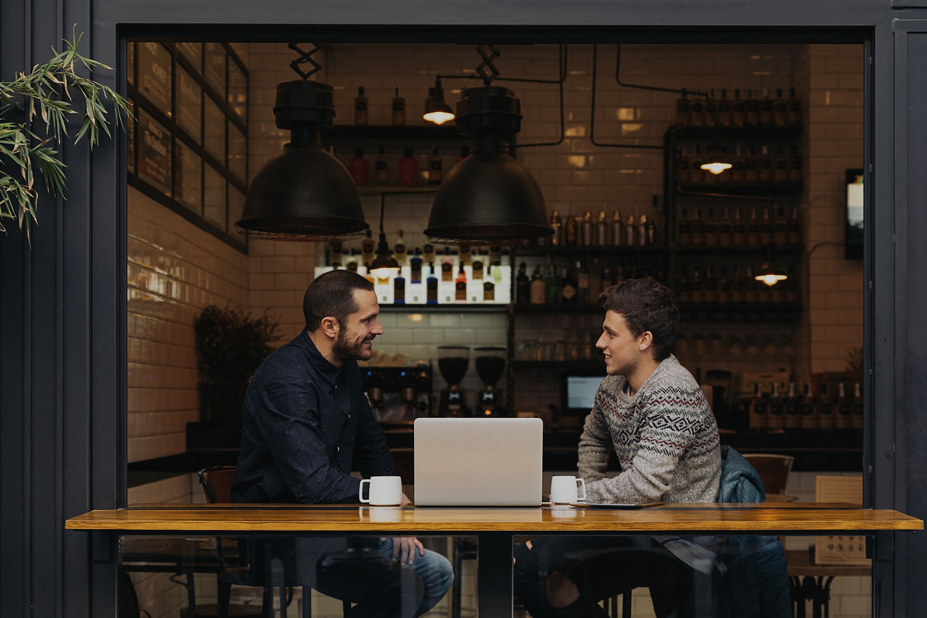 two men sat facing each other either side of a laptop in a modern coffee shop